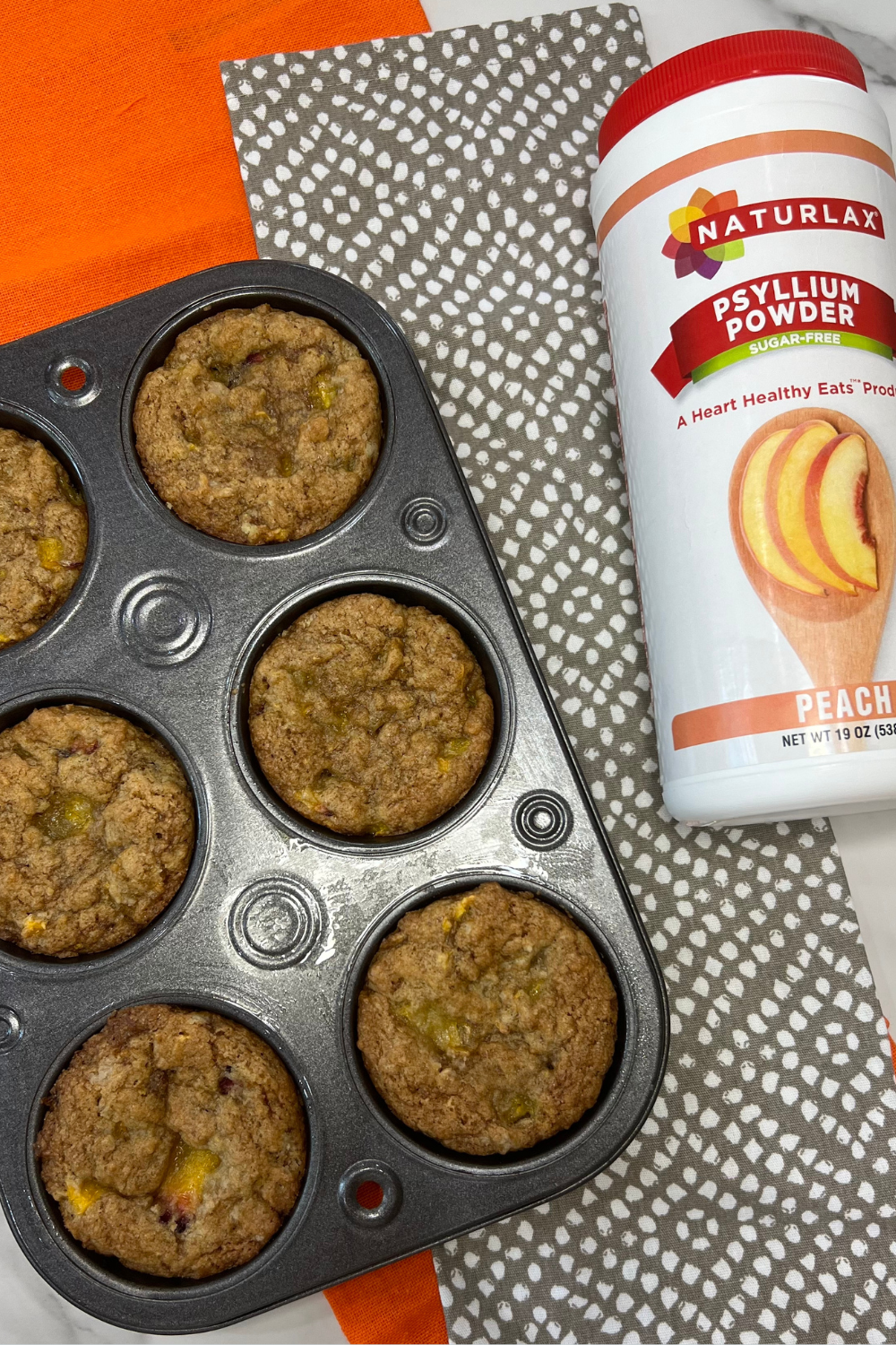 A muffin tray with six Easy One Bowl Peach Muffins next to a container of peach-flavored psyllium powder rests on a dotted cloth and an orange napkin.