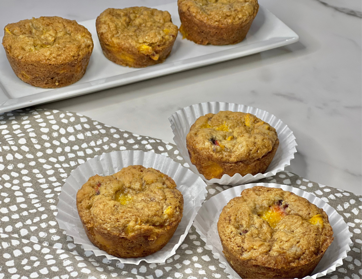 Four easy one bowl vegan peach muffins in paper liners are displayed on a white patterned cloth, with four more muffins on a rectangular white plate in the background. The scene is set on a marble countertop.