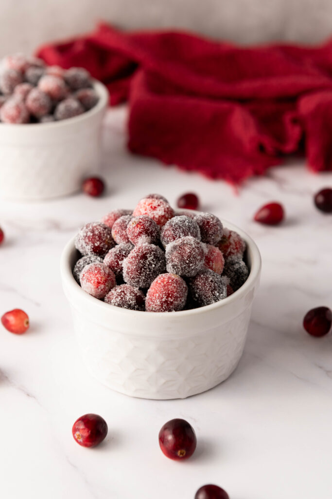 Two white bowls filled with sugared cranberries on a marble surface, surrounded by scattered cranberries, offer a tempting glimpse into how to make sugared cranberries. A red cloth adds a festive touch in the background.