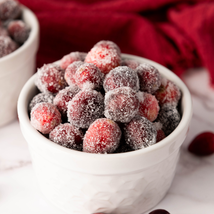 A white bowl brimming with delectable sugared cranberries rests on a marble surface, framed by a red cloth in the background. Perfect for those curious about how to make cranberries taste divine with just a dusting of sugar.
