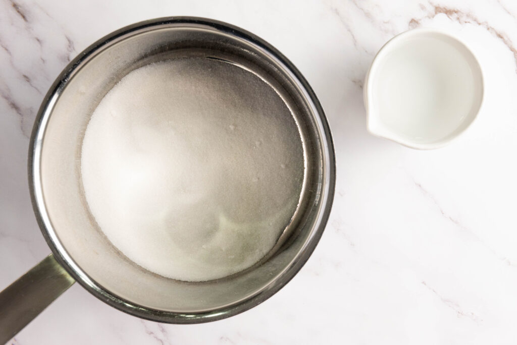 A pot filled with sugar next to a small white jug on a marble surface sets the scene for learning how to make sugared cranberries.