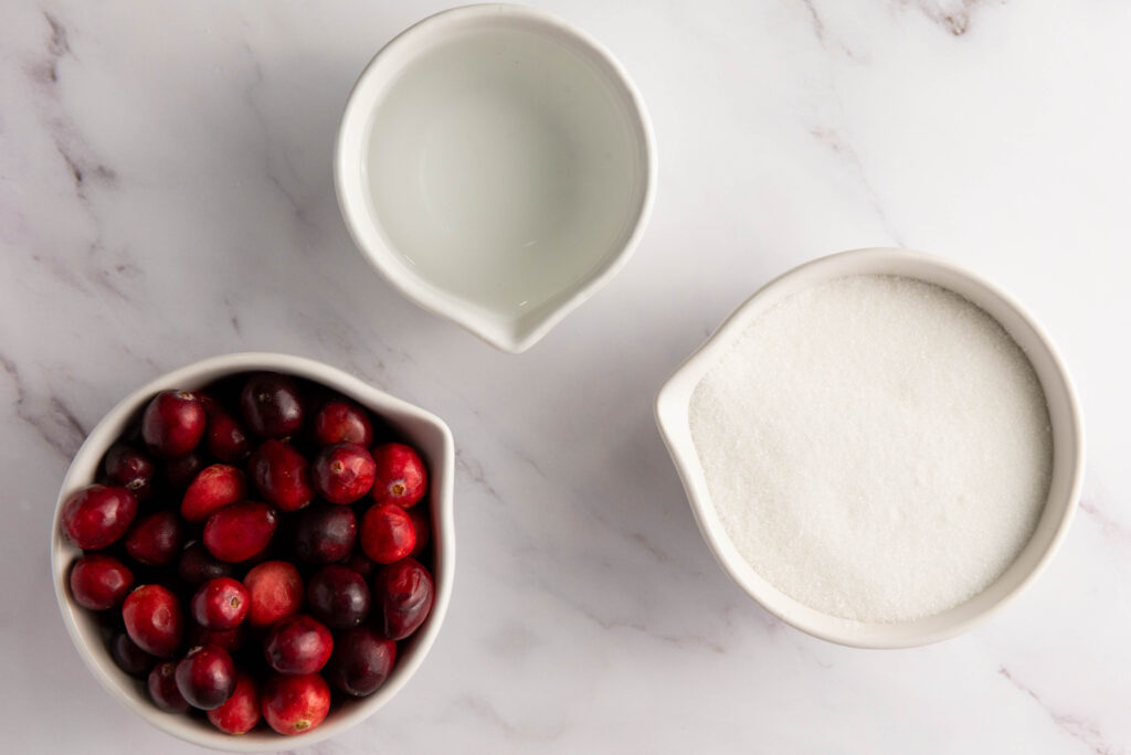 Three white bowls on a marble surface hold the essentials to make sugared cranberries, featuring vibrant cranberries, clear water, and fine sugar.
