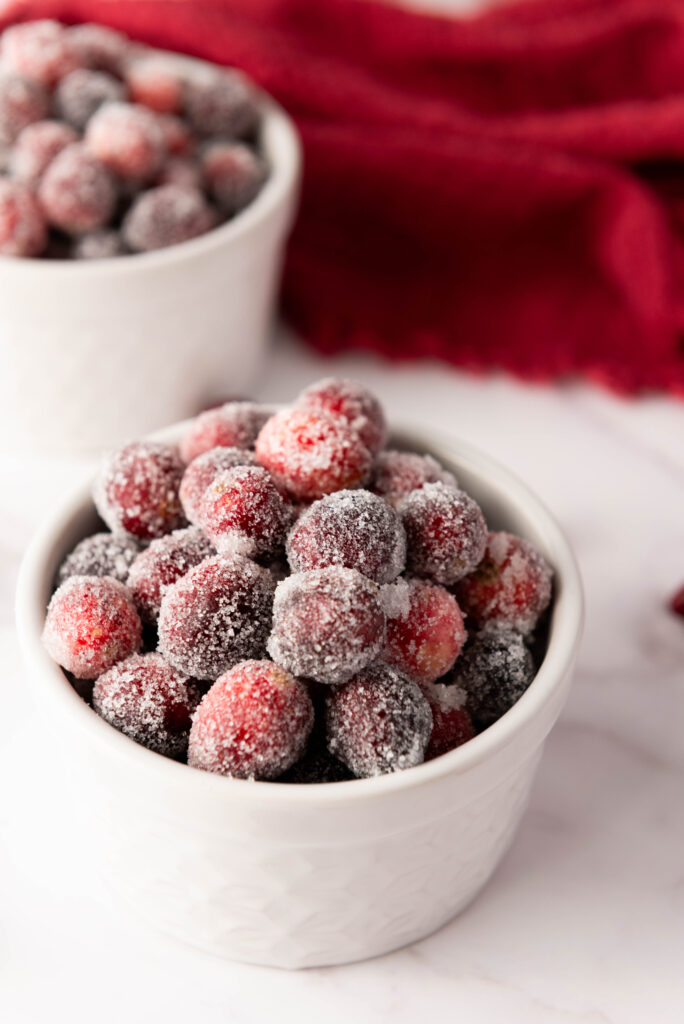 Two white bowls brimming with sugared cranberries sit elegantly on a white surface, accented by a red cloth in the background. Learn how to make these sweet treats that add a touch of festive charm to any setting.