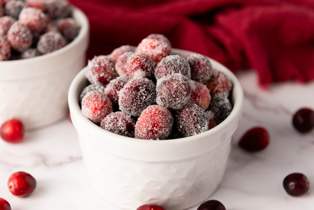 A white bowl filled with sugared cranberries sits on a marble surface, inviting you to discover a delightful recipe. A dark red cloth and more cranberries complement this sweet scene in the background.