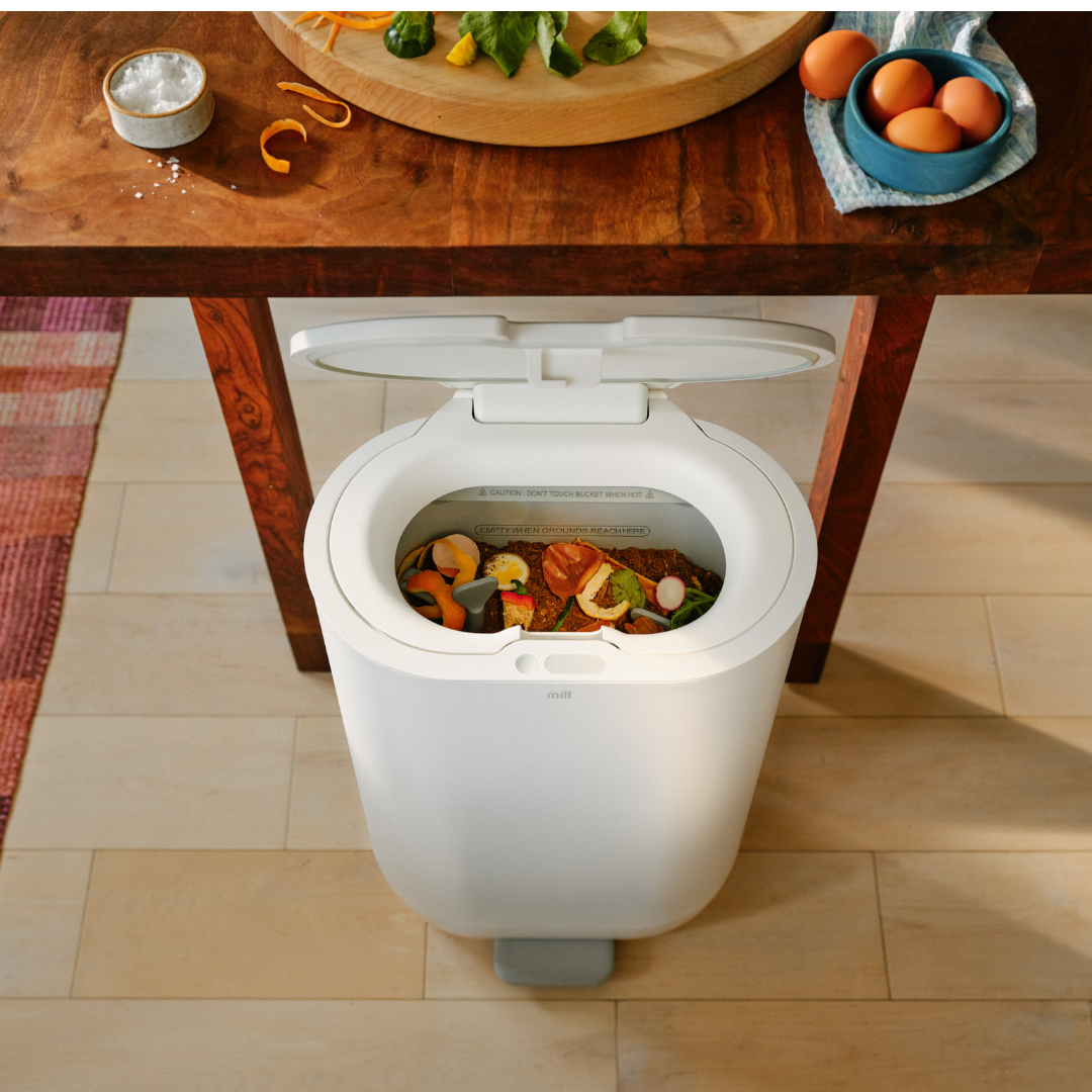 A white kitchen waste bin, resembling the efficiency of a Mill Food Recycler, sits with its lid open under a brown wooden table, revealing assorted food scraps like peels and vegetable pieces inside.