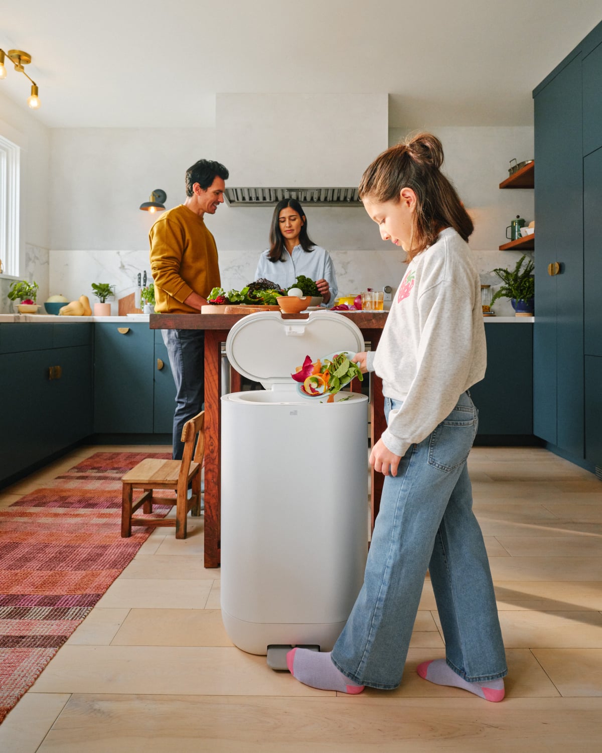 In a brightly lit kitchen, a child responsibly disposes of vegetable scraps in a Mill Food Recycler bin, while two adults prepare vegetables at the kitchen island.