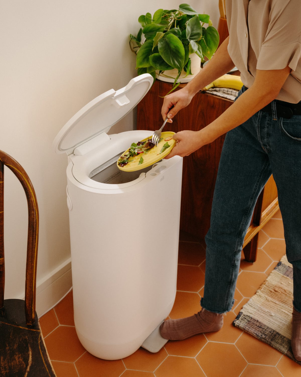 A person is thoughtfully disposing of food waste into a sleek white compost bin in their kitchen, with a lush plant standing nearby, thanks to their Mill Food Recycler.