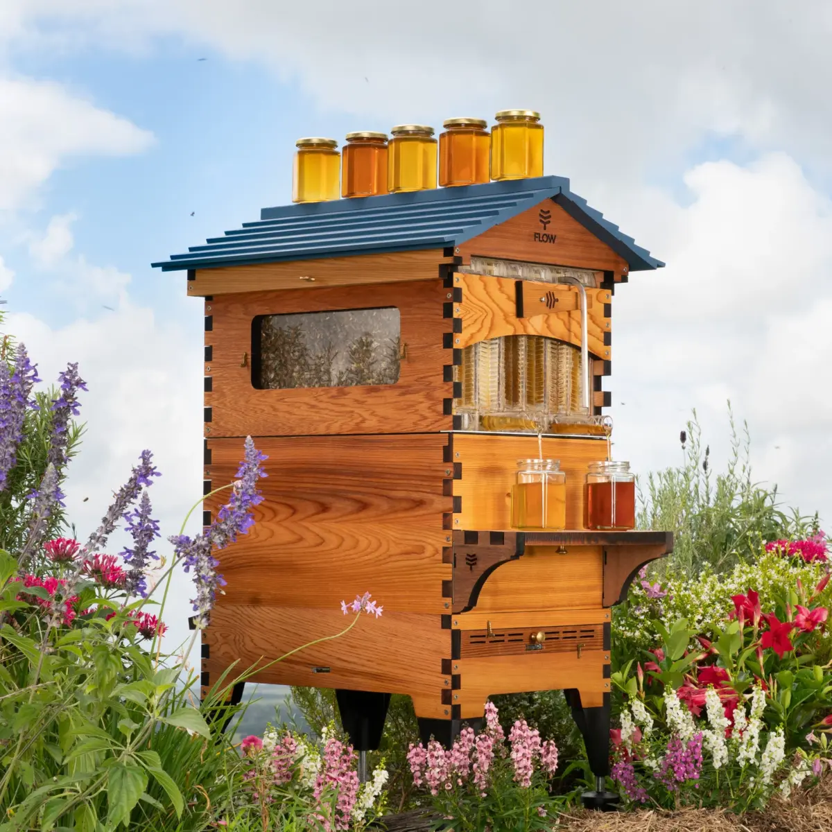 A wooden beehive with glass jars filled with honey sits gracefully among blooming plants, a testament to nature's harmony. Enjoy this serene setup and claim your Flow Hive Discount for a sweet deal on nature's finest treasure.