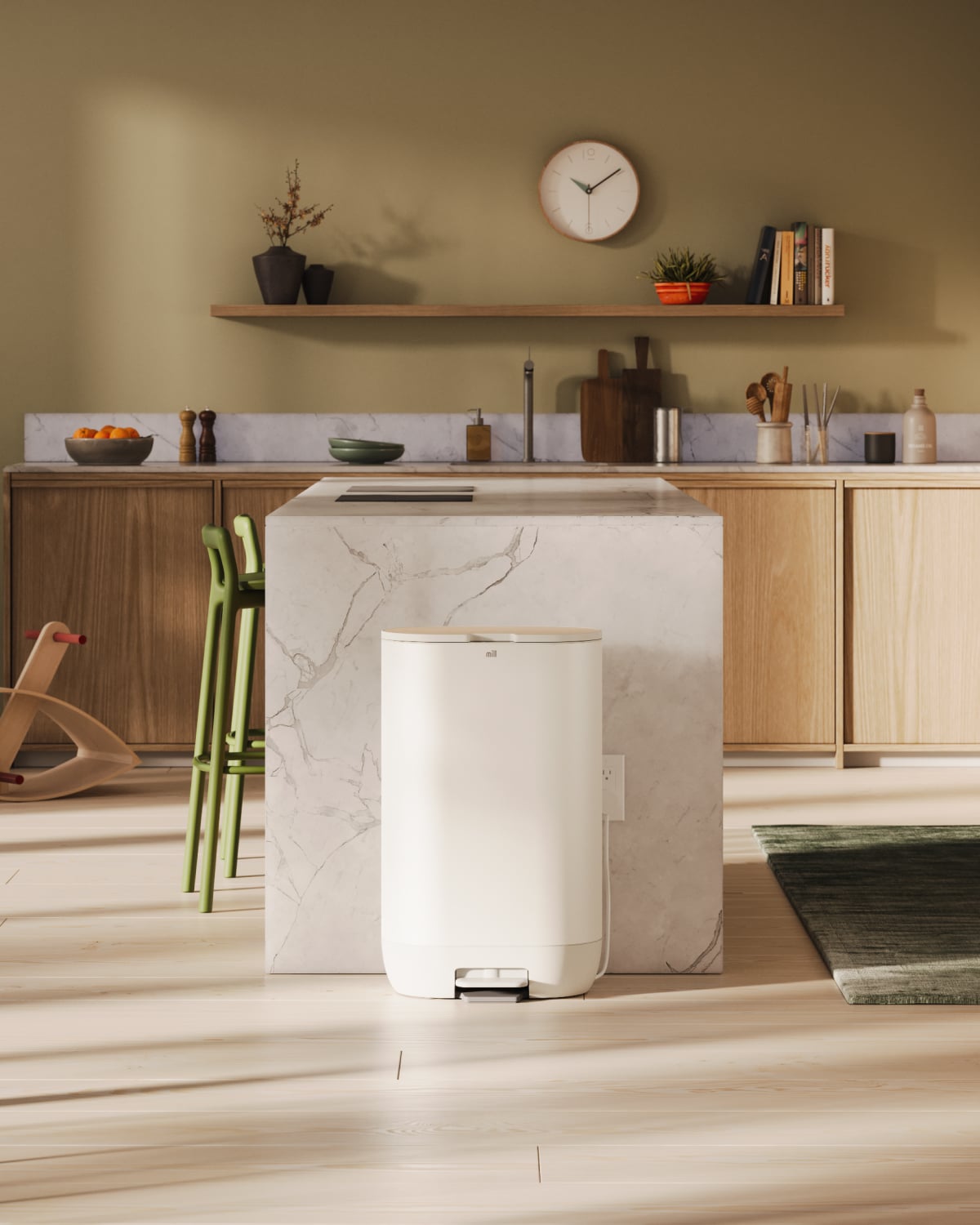 A modern kitchen features a marble island surrounded by green stools and wooden cabinets, complete with a white pedal bin perfect for food recycling. A green wall hosts a shelf adorned with plants, books, and a clock.