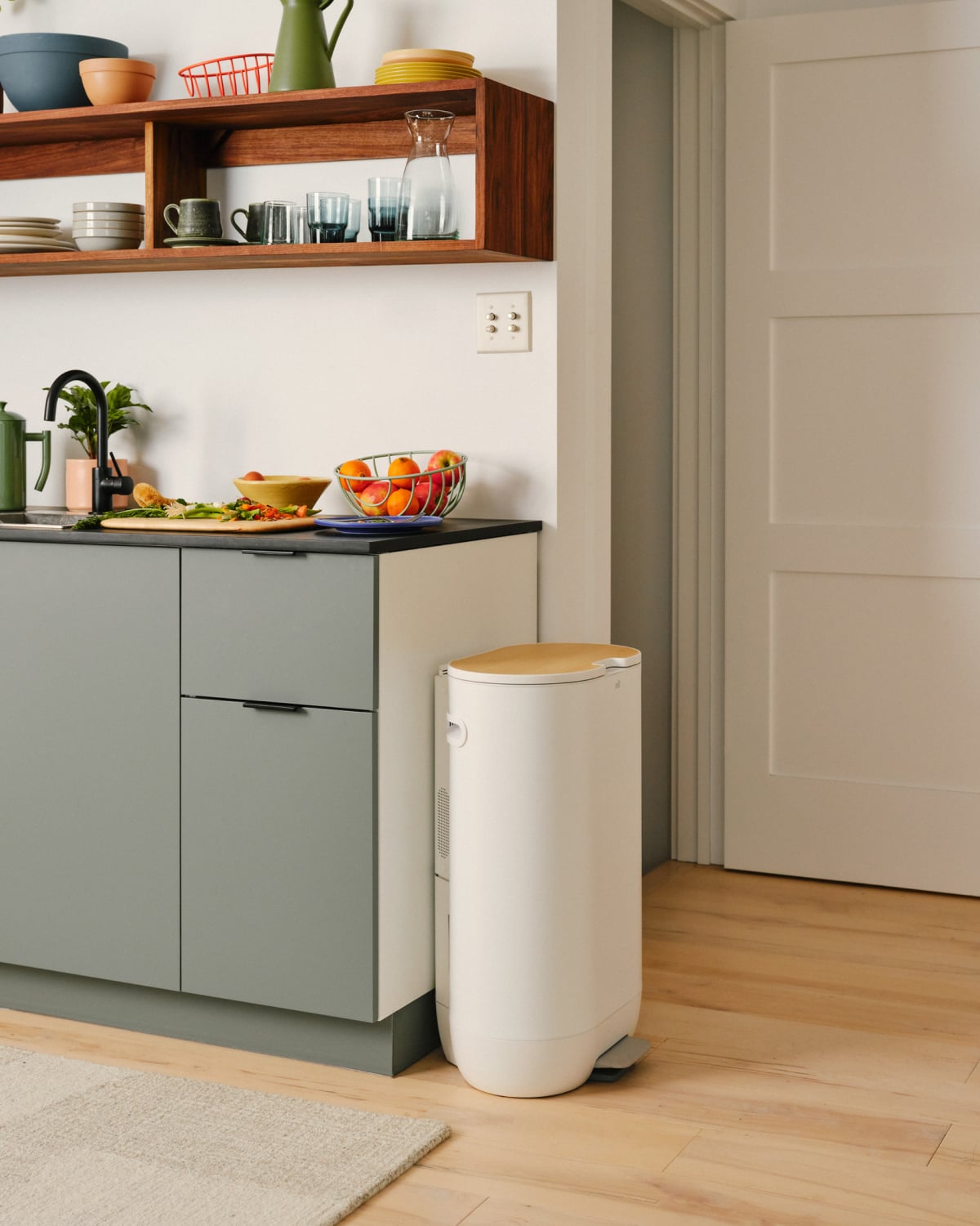 A modern kitchen at 10 Mill features grey cabinets, a black countertop, and open shelving. Near the doorway, you'll find a white trash bin with a wooden lid. A bowl of fruit graces the counter alongside a sleek food recycler that highlights the benefits of sustainable living.