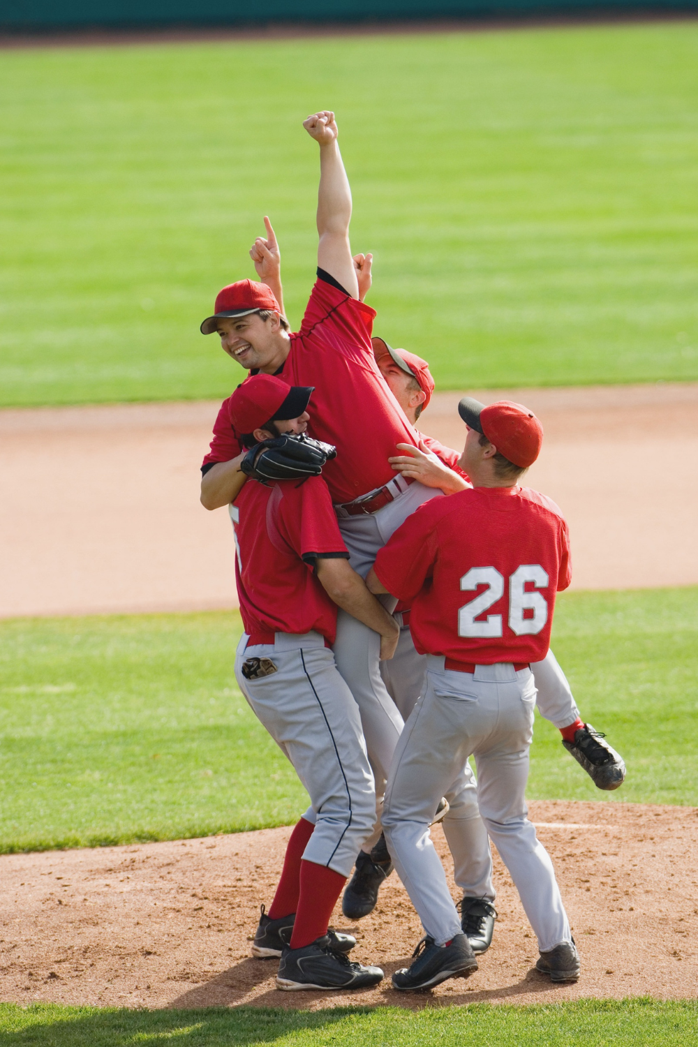 The BallBoyz Teens, a baseball team in red uniforms, jubilantly celebrate on the field, lifting a teammate who raises his arm triumphantly.