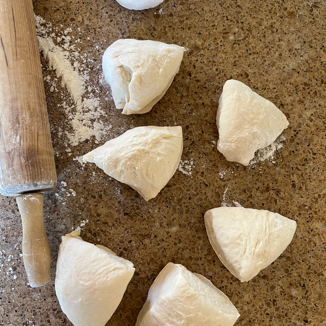 Several pieces of raw dough for an Easy Flatbread Recipe are arranged on a brown countertop near a wooden rolling pin, with some flour scattered around.