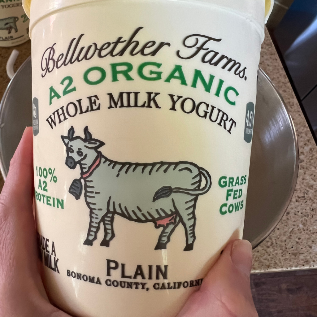 A person holds a container of Bellwether Farms A2 Organic Whole Milk Yogurt, featuring a cow illustration and product details—perfect for making delicious Yogurt Flatbread or any easy flatbread recipe.