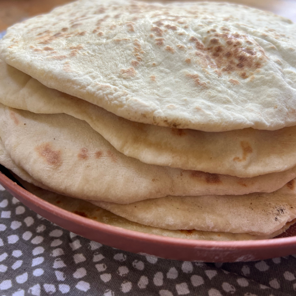 A stack of round, lightly browned yogurt flatbreads sits on a pink plate atop a patterned fabric surface.